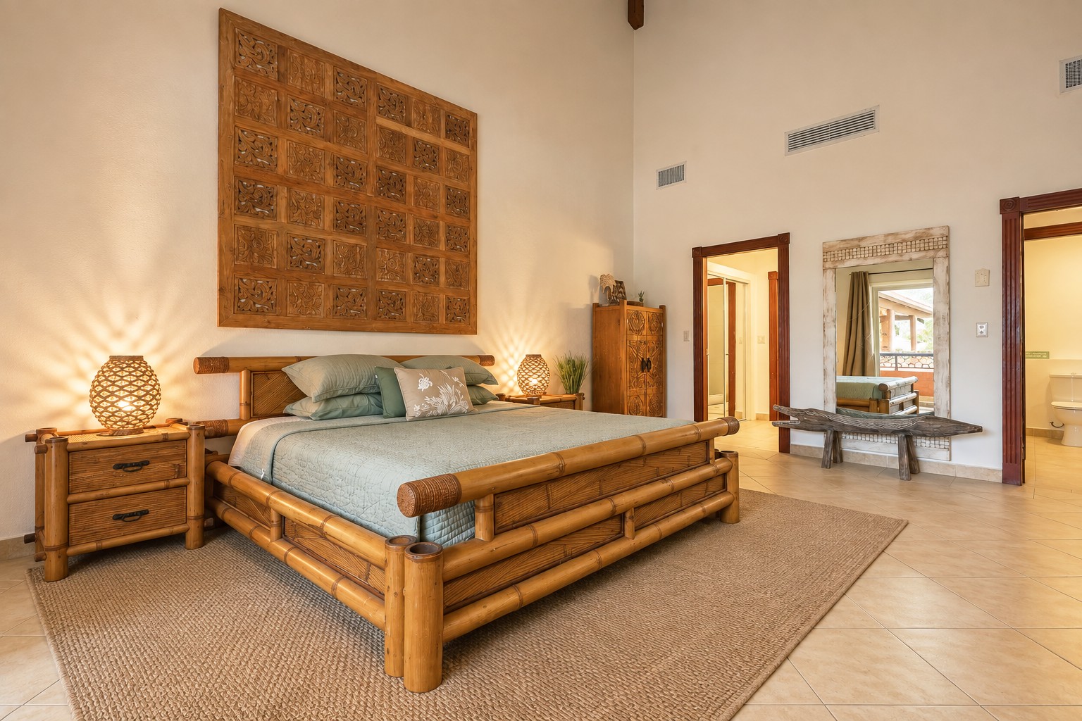 Warmly lit primary bedroom with a bamboo bed, sage bedding, carved wooden wall panel, woven lamps and a tall mirror reflecting the terrace.