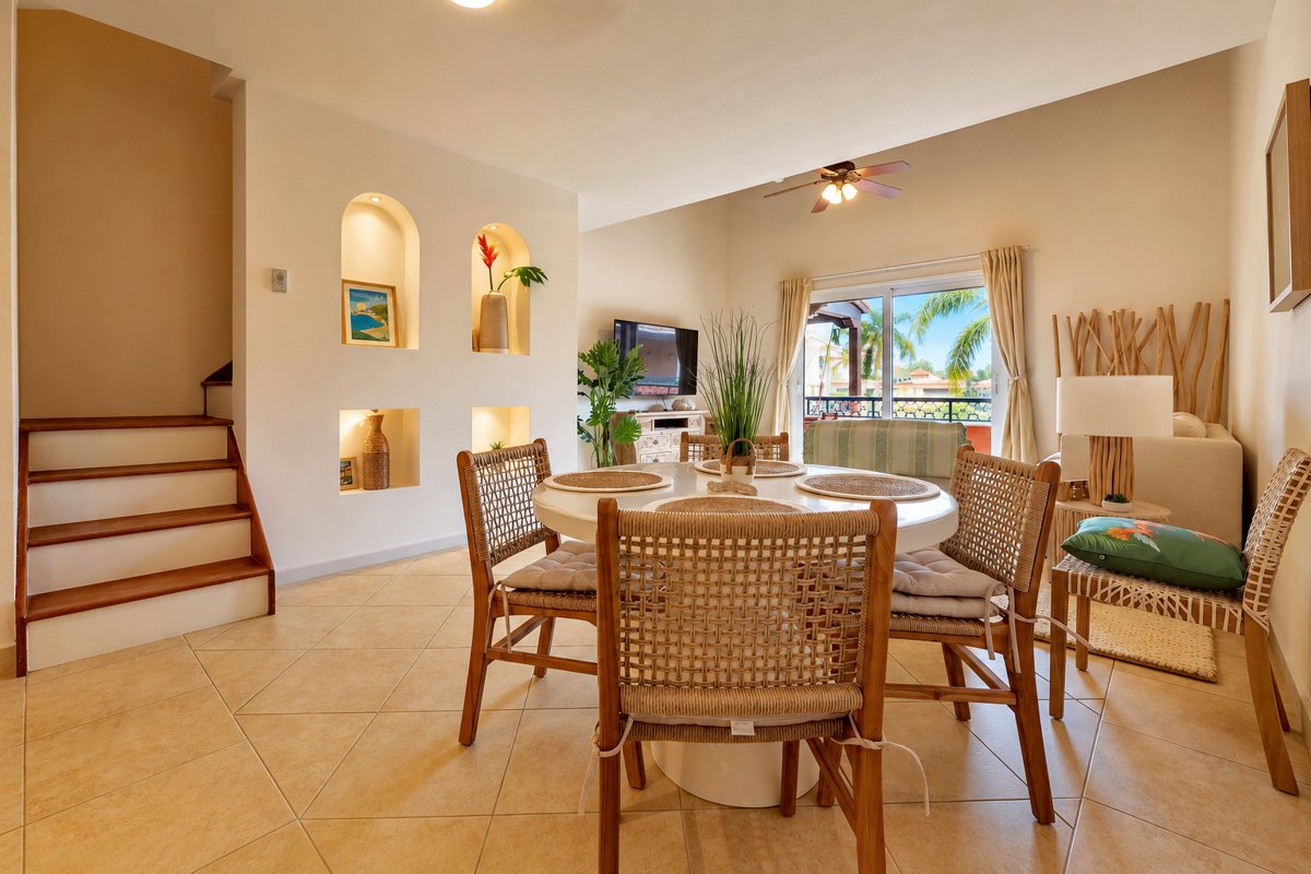 Sunlit dining area with a round table and woven chairs, illuminated arched niches, stairs to the loft, and the terrace beyond the living area.