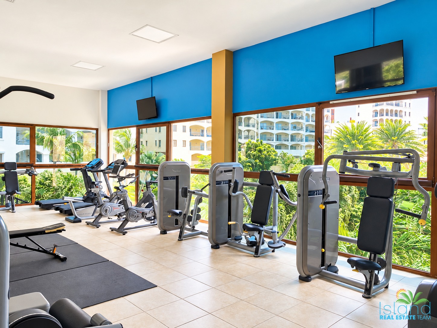 Strength and cardio equipment beside large gym windows overlooking tropical greenery.