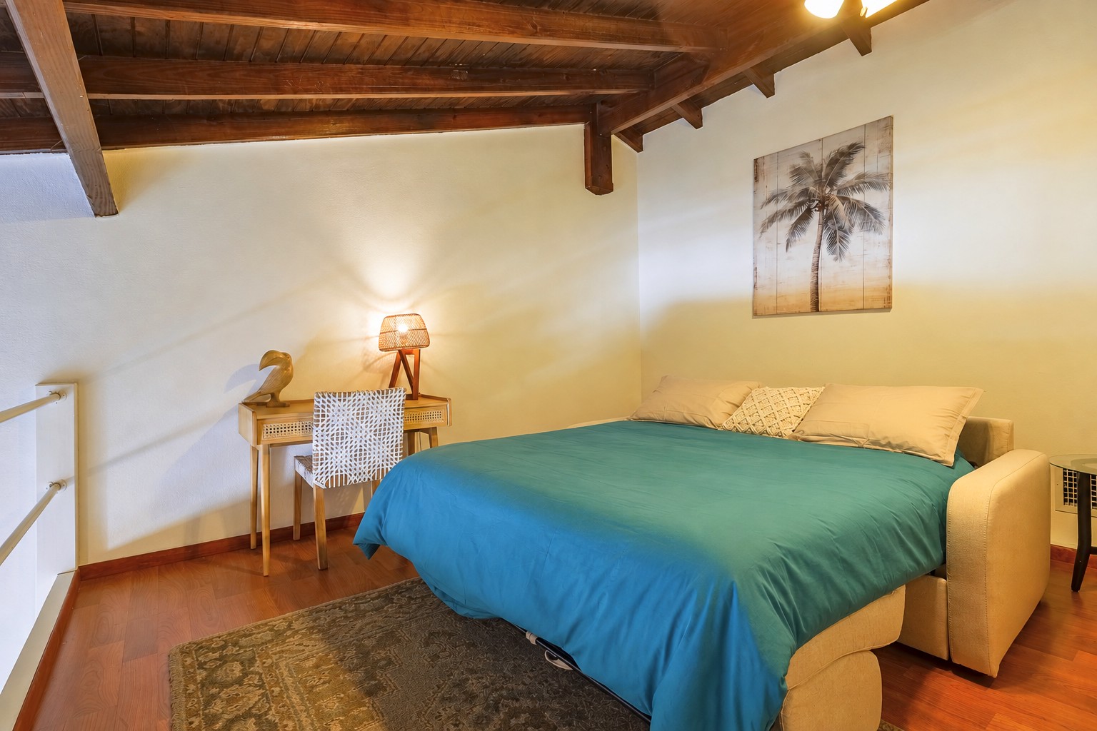 Queen sofa-bed prepared for sleep in the loft, with turquoise bedding, a desk and warm woven lamp.