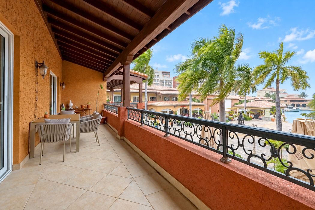 Covered marina-side terrace with a dining table and cushioned chairs, overlooking palm trees, the plaza and boats at Porto Cupecoy.